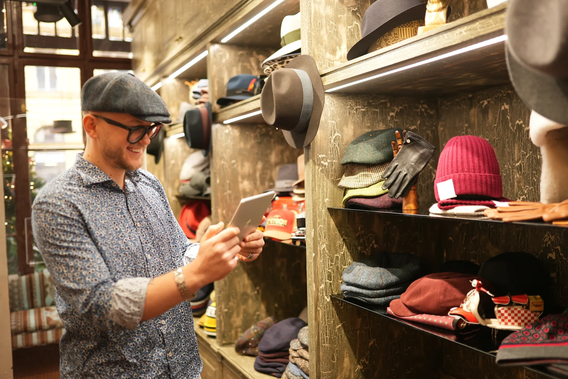 A shopkeeper in a hat shop using a tablet beside shelves of hats.